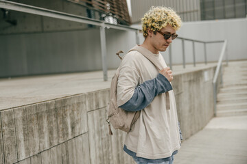 Fashionable young caucasian guy wearing backpack and sunglasses spending time outdoors. Blonde stylish man with stairs in background. Concept stylish weekend.