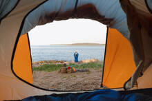 View From A Tent On The Shore Of A Lake Or Sea, A Woman Collects Firewood Or Does Yoga And Meditation. Camping In Nature, Wild Life In A Tent. Freedom And Relaxation From The Hustle And Bustle Of The