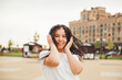 © inna717 - A young cheerful happy woman with dreadlocks, dressed in a white T-shirt, dancing, listening to music with headphones, resting, relaxing in a city park, walking along an alley. Urban lifestyle concept