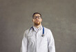 Portrait of a male doctor with a funny surprised and scared expression on a gray background. Caucasian man in glasses and a medical gown with a strange grimace looks into the camera.