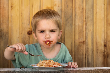 A little boy is eating spaghetti. His face is smeared with ketchup