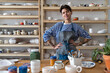 © DimaBerlin - Happy joyful young Turkish woman successful self-employed potter wearing apron behind table with pottery tools smiling at camera, cheerful female ceramist posing against display of handmade ceramics