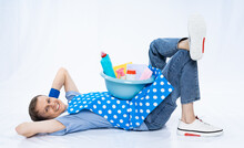 A Man In A Blue Apron Rubber Gloves A Blue T-shirt, Jeans And White Sneakers Lies On The Floor, On His Back, Put His Hands Under His Head. Put The Basin With Detergents On Himself. Isolated Background
