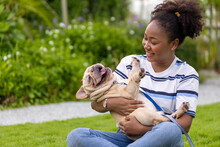 African American Woman Is Playing With Her French Bulldog Puppy While Walking In The Dog Park At Grass Lawn After Having Morning Exercise During The Summer