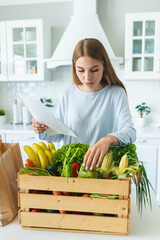 Woman take out fresh vegetables and fruits from wooden box on table in kitchen carefully checking on list. Organic products without nitrates. Vegetarianism, nutrition, healthy eating, cooking.