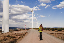 Technician With Rope Standing On Road Looking At Wind Turbine