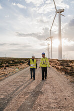 Technician And Colleague Discussing Walking On Dirt Road At Wind Turbines