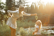Side view of family standing near pond in park lit up by sunset in summer. Young woman holding grapes over little girl.