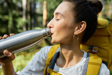 Close Up Of Thirsty Woman Backpacker Drinking Fresh Water Or Tea During Stop In Trekking In Wild Nature, Enjoying Drink With Closed Eyes, Carrying Rucksack On Her Shoulders With Forest On Background