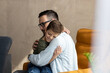 © Panorama - A loving parent sits on the sofa at home and hugging his son. The child is sitting in fathers lap and enjoying moment with his father