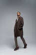 full length of young african american man in autumnal coat looking at camera and posing with hand in pocket on grey.