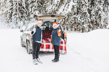 Wall Mural - Two friends teenagers boys in red santa hats with with sparkles Bengal fire standing near trunk of car decorated for Christmas and New Year in snowy winter forest. Road trip and local travel.