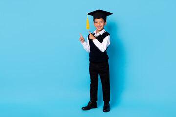 Full length photo of charming funny small boy wear black waistcoat graduation cap pointing empty space isolated blue color background