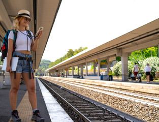 Woman waits for train at station while working happily on phone, digital nomad, travel and transportation concept