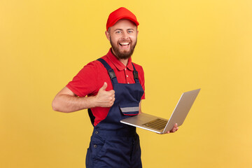 Wall Mural - Satisfied worker man wearing blue overalls, red T-shirt and cap working on laptop, showing thumb up, likes a new online service, looking at camera. Indoor studio shot isolated on yellow background.