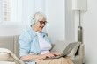 © Tatiana - a happy, joyful old lady is sitting on a beige sofa with glasses on her face and headphones on her head working at home in a comfortable environment learning new things