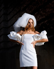 Cropped of elegant woman in wedding dress with veil in hair, puffy short sleeves and open neckline, holding hands on slim waist and posing outdoors while looking at sun