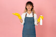 Attractive young Asian woman wearing apron with yellow rubber gloves and holding clean spray on pink background.
