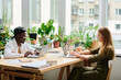 © pressmaster - Two young multicultural coworkers sitting by desk in front of laptops and networking against window in office with many green plants