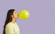 Profile portrait of young woman inflates yellow balloon on pastel purple background. Funny young ethnic brunette woman holding big chewing gum standing near copy space. Banner. Side view.