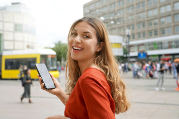 Wall Mural - Portrait of natural beautiful smiling woman walking in Berlin Alexanderplatz holding a mobile phone with blurred city background