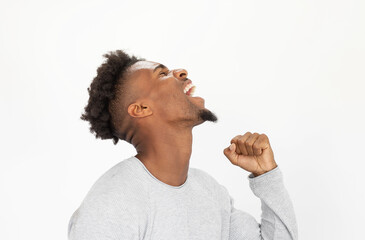 Wall Mural - Portrait of excited African American man wearing white sweater. Young bearded guy screaming and showing fist against white background. Success concept