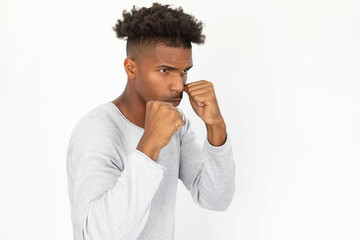 Wall Mural - Confident African American man standing in boxing stance. Portrait of young bearded guy wearing white sweater clenching fists and looking away against white background. Conflict and defense concept