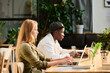 © pressmaster - Side view of two young intercultural designers or analysts networking by desk in front of laptop screens in openspace office with green plants