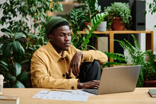 Young Serious Black Man In Casualwear Sitting By Workplace In Front Of Laptop Screen In Green Office And Working Over Business Project
