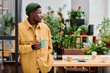 © pressmaster - Young black man in stylish casualwear holding cup of coffee while standing in front of camera against his workplace in office