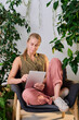 © pressmaster - Serious blond female manager in casualwear scrolling in tablet while sitting on chair among green plants in modern office