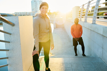 Wall Mural - Happy young blond sportswoman in activewear standing on bridge by riverside against sunlight and African American athlete