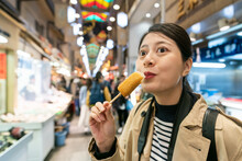 Portrait Of Amazed Asian Chinese Girl Visitor Eating Yummy Oden Stick With Satisfied Look On Face On Teramachi Dori In Nishiki Market In Kyoto Japan