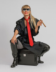 Wall Mural - young blonde woman with long hair in sunglasses with a cigar in her hand sits on a suitcase on a white background