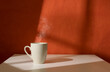 Cup of morning fragrant coffee in rays of sunlight from window. White cup with steaming coffee on white table.