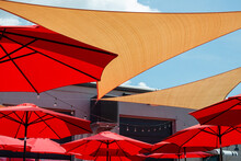 Multiple Triangle Shaped Yellow Nylon Sunshades And Awnings Hanging Over A Patio Deck. There Are Red Colored Canvas Umbrellas Hung With Strings Of Clear Patio Light Against A Bright Blue Sunny Sky.