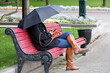 Rain in a city, woman wearing jeans and brown boots with sitting with umbrella on bench and using smartphone. Rainy weather in autumn park