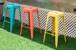 Three colorful iron metal stackable bar stools, blue, peach, and yellow color. The chairs are on green grass with white lattice in the background. The summer outdoor garden has shade and sunny spots.