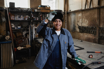 Waist up portrait of multiethnic female welder smiling at camera in industrial factory workshop, copy space