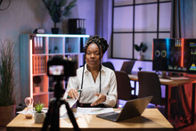 Cheerful Confident African American Businesswoman, Broker In White Shirt Sitting In Front Of Camera In Evening Office During Recording Video For Business Vlog.