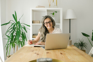 Portrait of adult woman working from home and looking at the camera, she is sitting at her desk with laptop open next to her and smiling to camera