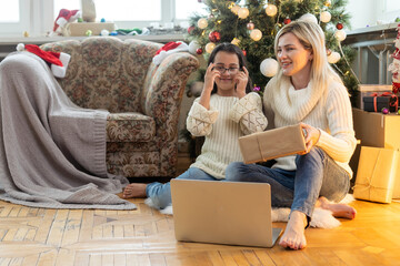 Wall Mural - Indoor shot of beautiful happy young woman shopping online on laptop in cozy Christmas interior. Mother on the floor next the Christmas tree and sofa and daughter embrace her, shopping gifts.