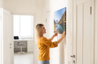 © Angelov - Modern Home Interior And Domestic Decor. Smiling young woman hanging painting, putting photo picture frame on the wall. Casual lady taking care of coziness in her new stylish apartment, profile