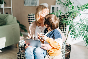 Senior woman and son kid playing with tablet pc. Smiling woman and her grandson sitting home using gadget digital device for online shopping internet surfing video conference calling.