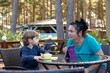 © Татьяна Волкова - Little boy sit on chair at table on summer terrace and talk with mother. Kid of kindergarten age having dinner outdoor in forest. Communication with people, health resort, vacation, camp in forest