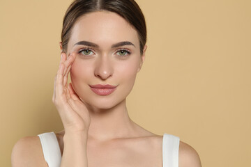 Poster - Young woman applying cream under eyes on beige background, space for text