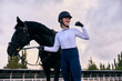 Winner. Young sportive girl, professional jockey or horsewoman in sports uniform and helmet with black horse at riding arena. Horseback riding