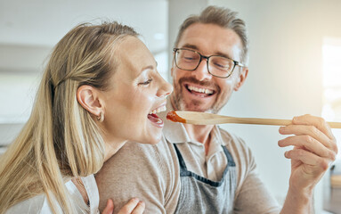 Wall Mural - Happy couple cooking, tasting spoon of food and meal, lunch and dinner in kitchen at home. Smile mature man giving hungry woman a taste of homemade tomato sauce meal in mouth to enjoy eating together