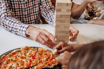 A group of multiage and multiethnic people playing jenga game on a table at home,hands close up.People eat pizza while playing.Board games concept.Time together.Stay home concept.Selective focus.