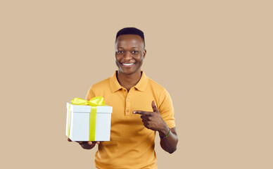 Happy handsome young man holding present tied with ribbon. Cheerful attractive African American guy in casual yellow T shirt smiling and pointing at white gift box with yellow bow in his hand